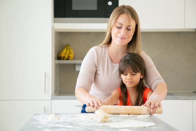 Joyeuse maman et sa fille cuisiner ensemble, rouler la pâte sur la table de cuisine avec de la farine en poudre.