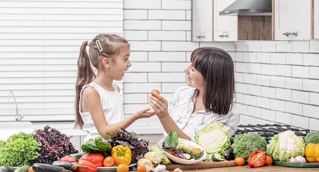 Joyeuse maman et sa fille préparent une salade de légumes à partir de nombreux légumes dans une cuisine moderne et légère.