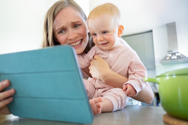 Joyeuse maman et sa petite fille riant à l'écran de la tablette, regardant un séminaire en ligne ou ayant un appel vidéo. Concept de garde d'enfants ou de cuisine à la maison