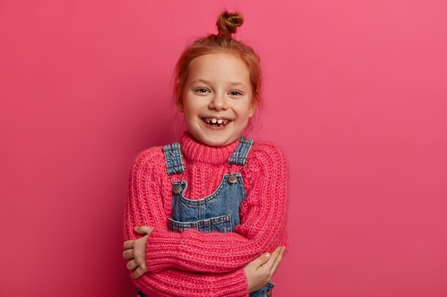 Joyeuse petite fille au gingembre se serre dans ses bras, se sent à l'aise, a un nouveau pull rose en laine, une tenue douce et chaude, sourit à pleines dents, montre des dents manquantes, a les cheveux roux, isolés sur un mur rose.