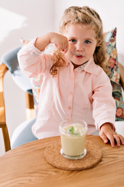 Joyeuse petite fille aux cheveux bouclés portant une chemise rose mangeant un smothie vert au café