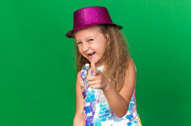 joyeuse petite fille blonde avec un chapeau de fête violet pointant isolé sur un mur vert avec espace pour copie