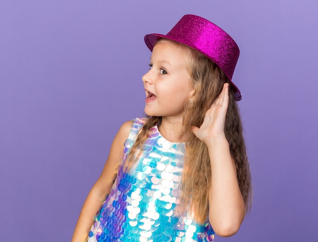 Joyeuse Petite Fille Blonde Avec Un Chapeau De Fête Violet Tenant La Main Près De L'oreille Essayant D'entendre Isolé Sur Un Mur Violet Avec Espace De Copie