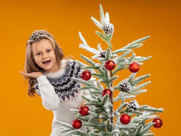 Photo gratuite joyeuse petite fille debout derrière l'arbre de noël portant diadème avec guirlande sur le cou propagation main isolé sur fond orange