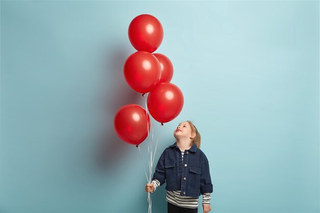 Joyeuse petite fille lève la tête et regarde attentivement les ballons à air rouge, porte une veste à la mode en denim, se prépare à célébrer son anniversaire, des modèles sur un mur bleu, joue à l'intérieur. Fête pour les enfants