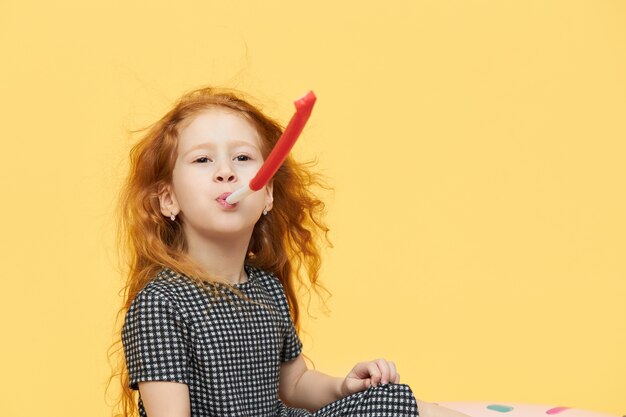 Joyeuse petite fille ludique aux longs cheveux roux soufflant la corne de fête