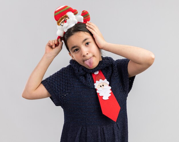 Joyeuse petite fille en robe en tricot portant une cravate rouge avec jante de Noël drôle sur la tête s'amusant à sortir la langue