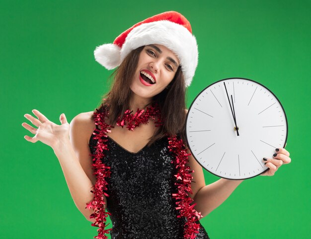 Joyeuse tête inclinable jeune belle fille portant un chapeau de Noël avec guirlande sur le cou tenant horloge murale propagation main isolé sur mur vert