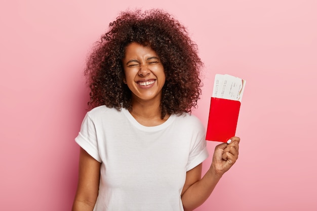 Photo gratuite joyeuse touriste s'amuse avant le voyage, détient les billets d'embarquement dans le passeport, se sent excitée par le voyage, sourit positivement, porte une tenue décontractée, isolée sur un mur rose, attend le vol