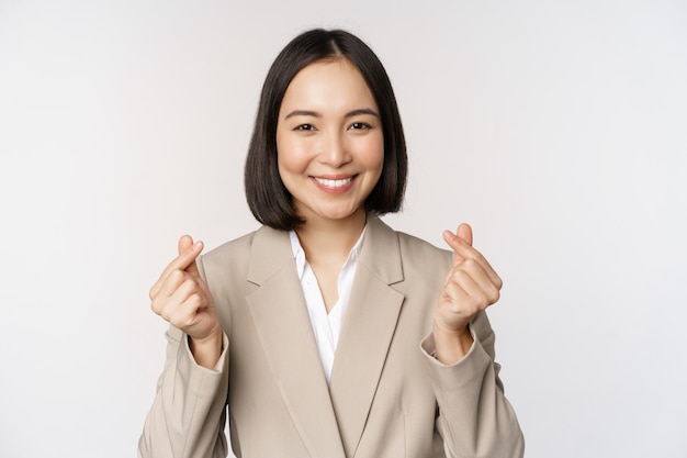 Joyeuse vendeuse asiatique souriante et montrant un signe de coeur de doigt debout en costume sur fond blanc