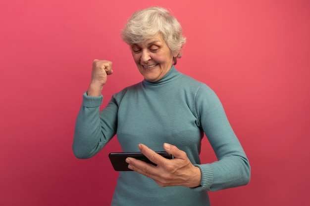 Joyeuse vieille femme portant un pull à col roulé bleu tenant et regardant un téléphone portable faisant un geste oui