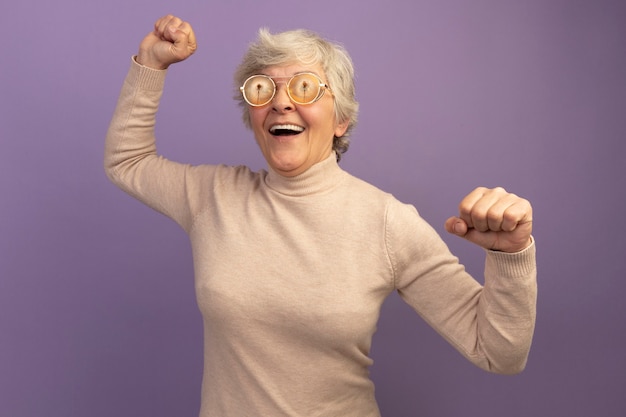 Joyeuse vieille femme portant un pull à col roulé crémeux et des lunettes regardant le côté levant les poings isolés sur un mur violet