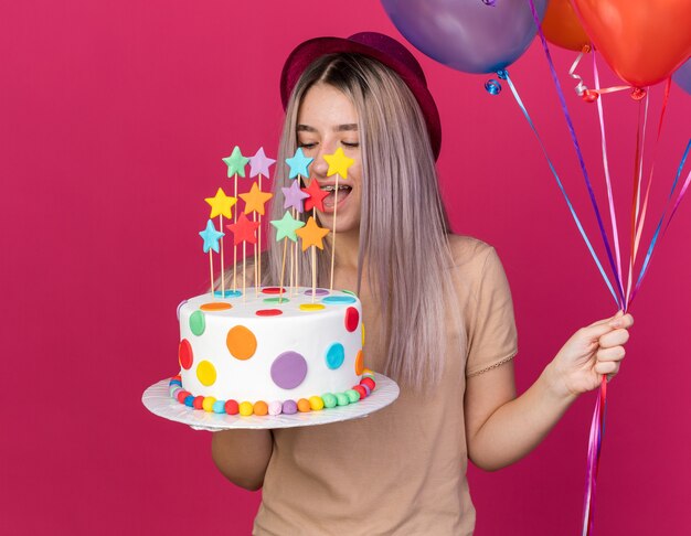 Joyeuse avec les yeux fermés jeune belle fille portant un chapeau de fête tenant des ballons avec un gâteau