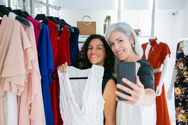 Joyeuses amies mignonnes appréciant le shopping dans la boutique ensemble, tenant la robe, s'amusant et prenant selfie sur téléphone mobile. Concept de consommation ou d'achat