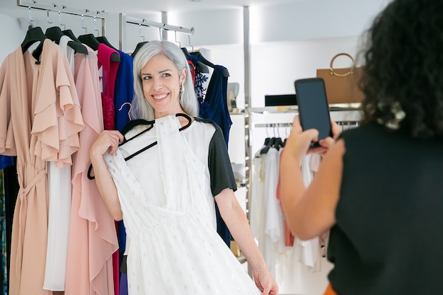 Joyeuses dames appréciant le shopping dans un magasin de mode ensemble, tenant une robe et prenant des photos sur téléphone portable. Concept de consommation ou d'achat