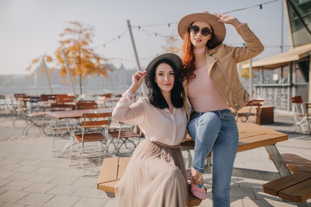 Joyeuses Femmes Touchant Des Chapeaux Tout En Posant Dans Un Café De Rue