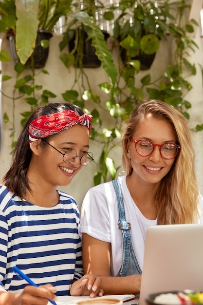 Joyeuses filles brune et blonde utilisent un ordinateur portable moderne pour se divertir et étudier, passer du temps libre ensemble