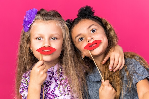 Photo gratuite joyeuses petites filles jolies tenant de fausses lèvres sur des bâtons isolés sur un mur rose avec espace de copie