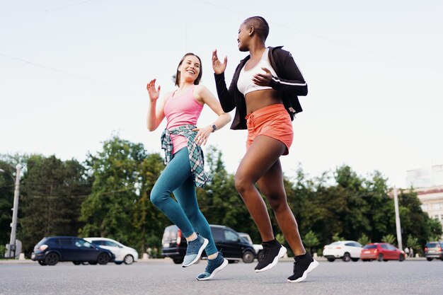 Joyeux amis souriants en vêtements de sport courant dans la ville discutant des femmes multiethniques ayant un entraînement de fitness