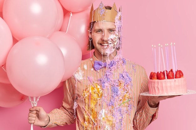 Joyeux anniversaire homme avec une expression heureuse porte des vêtements de fête sales couronne de papier tient le gâteau et les ballons pose à la fête contre le mur rose célèbre l'anniversaire ou l'obtention d'une nouvelle position