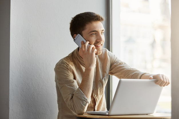 Joyeux beau brunet guy aux yeux gris travaillant dans la cafétéria sur ordinateur portable, souriant et parlant au téléphone avec le client sur les détails de la commande.