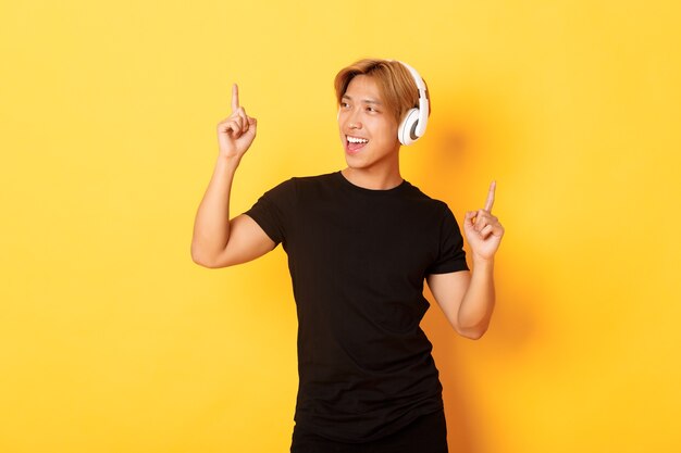 Joyeux beau mec asiatique aux cheveux blonds, chantant et dansant en écoutant de la musique dans des écouteurs sans fil, mur jaune debout