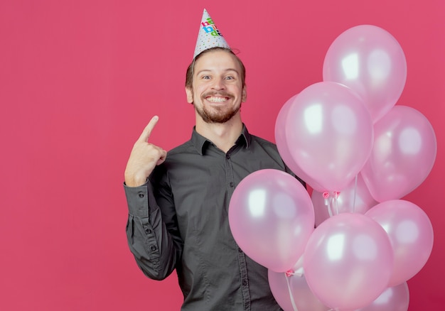 Joyeux bel homme au chapeau d'anniversaire se dresse avec des ballons d'hélium pointant sur le cap isolé sur le mur rose