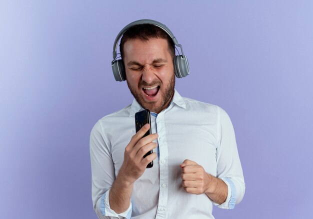 Joyeux bel homme sur le casque tient le téléphone faisant semblant de chanter isolé sur mur violet