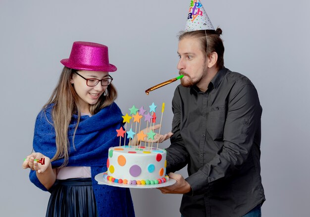 Joyeux bel homme en casquette d'anniversaire tenant un gâteau et sifflant en regardant une jeune fille surprise avec des lunettes portant un chapeau rose tient un sifflet et regarde un gâteau