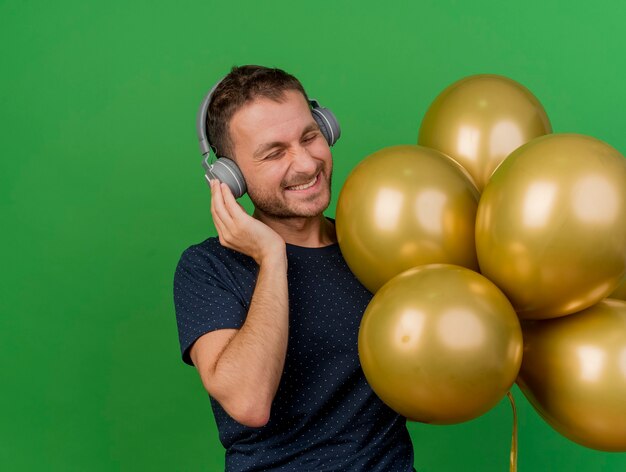 Joyeux bel homme caucasien sur les écouteurs détient des ballons d'hélium isolés sur fond vert avec espace copie