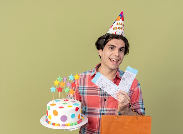 Joyeux bel homme caucasien portant une casquette d'anniversaire détient des billets d'avion en papier et un gâteau d'anniversaire