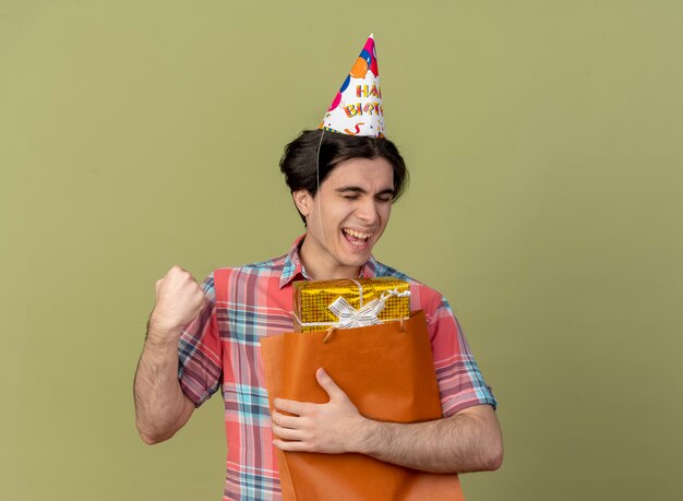 Joyeux bel homme caucasien portant une casquette d'anniversaire garde le poing et tient une boîte-cadeau dans un sac à provisions en papier