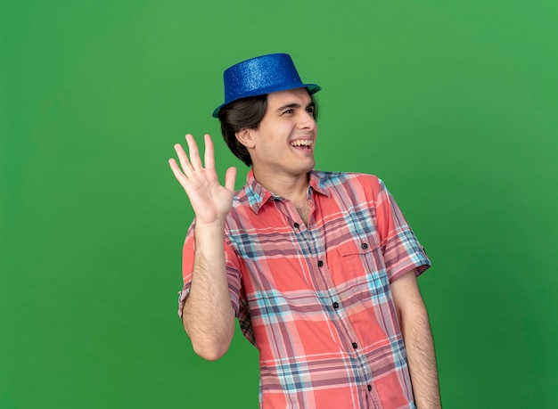 Joyeux bel homme caucasien portant un chapeau de fête bleu se dresse avec la main levée regardant de côté