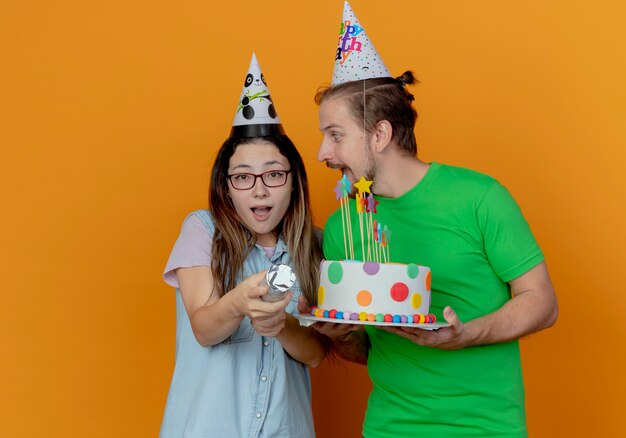 Joyeux bel homme en chapeau de fête tient le gâteau d'anniversaire et regarde surpris jeune fille portant chapeau de fête détient canon à confettis isolé sur mur orange