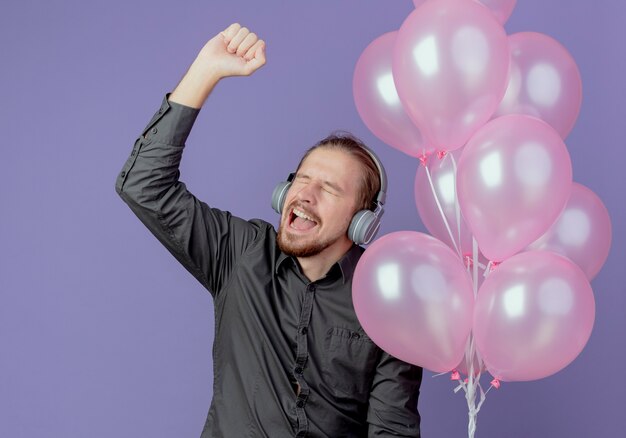 Joyeux bel homme sur les écouteurs détient des ballons d'hélium et lève le poing isolé sur mur violet