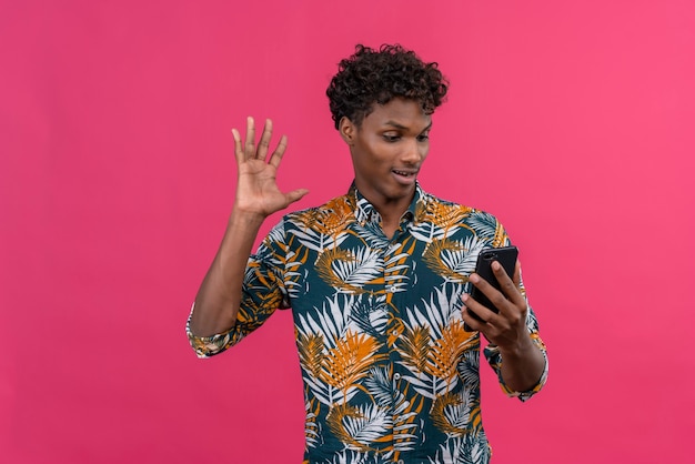 Joyeux bel homme à la peau sombre avec des cheveux bouclés en chemise imprimée de feuilles de parler avec un ami pendant un appel vidéo et en agitant la main sur la caméra avant du téléphone mobile sur fond rose