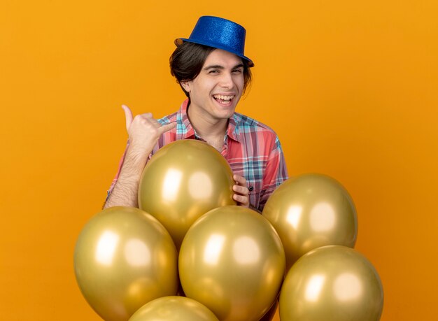 Joyeux bel homme portant un chapeau de fête bleu se dresse avec des ballons d'hélium faisant des gestes appelez-moi signe isolé sur mur orange