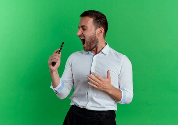 Joyeux bel homme tient le téléphone et regarde le côté faisant semblant de chanter isolé sur mur vert