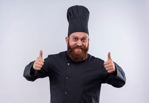 Un joyeux chef barbu en uniforme noir montrant les pouces vers le haut tout en regardant sur un mur blanc