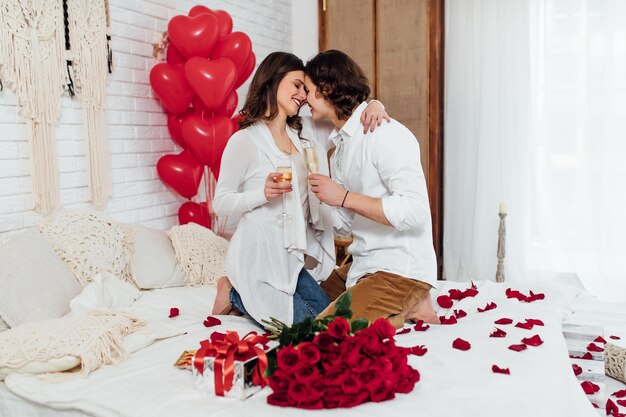 Joyeux couple assis sur le lit face à face avec des verres de champagne, célébrant ensemble la Saint-Valentin