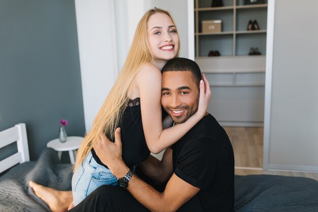 Joyeux couple heureux de jolie jeune femme aux longs cheveux blonds et joyeux beau mec étreignant sur le lit dans un appartement moderne. Joie, bonheur, sourire, matin ensemble, amour, loisirs, maison