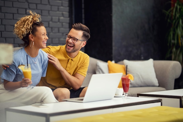 Joyeux couple riant tout en parlant de quelque chose de drôle et en utilisant un ordinateur portable dans un café