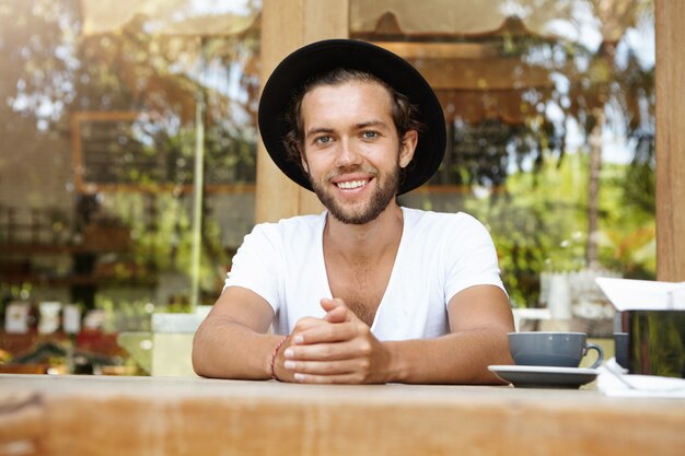 Joyeux étudiant bronzé avec une barbe épaisse ayant un bon café pendant le déjeuner, souriant joyeusement, profitant des vacances d'été dans un pays tropical