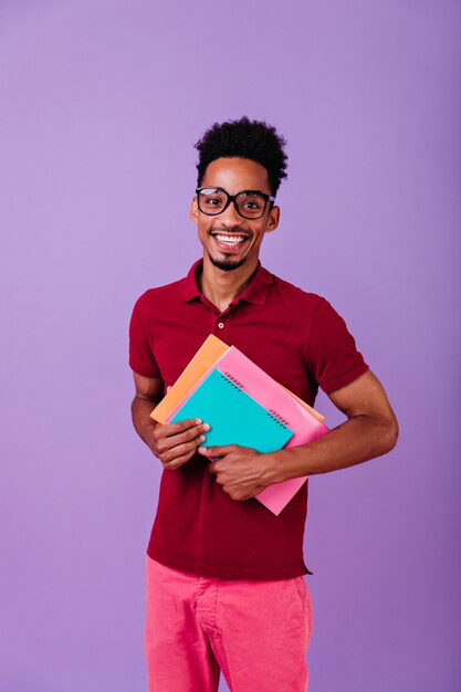 Joyeux étudiant international tenant des manuels colorés. Photo intérieure d'un homme africain en riant dans des verres isolés.