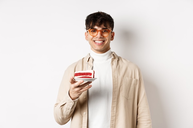 Joyeux gars de race blanche célébrant son anniversaire, tenant un gâteau avec une bougie et souriant gai à la caméra, debout sur fond blanc