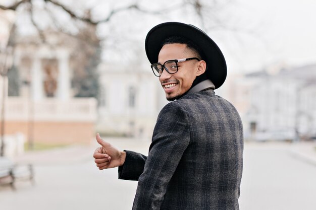 Joyeux homme africain regardant par-dessus l'épaule. Mec noir drôle en veste élégante montrant le pouce vers le haut avec le sourire.