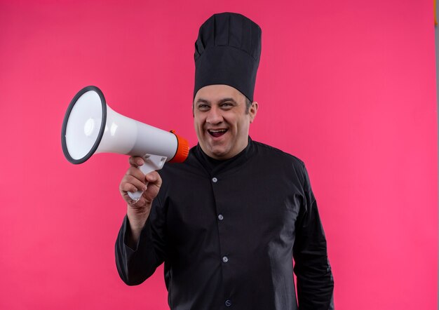 Joyeux homme d'âge moyen cuisinier en uniforme de chef parler par haut-parleur sur mur rose isolé