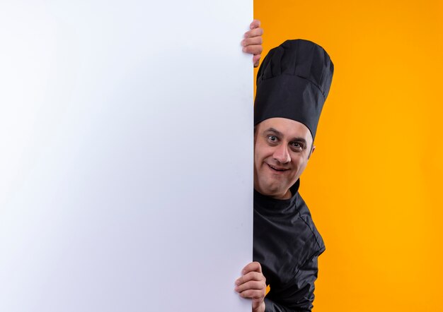 Joyeux homme d'âge moyen cuisinier en uniforme de chef tenant un mur blanc sur un mur jaune avec copie espace