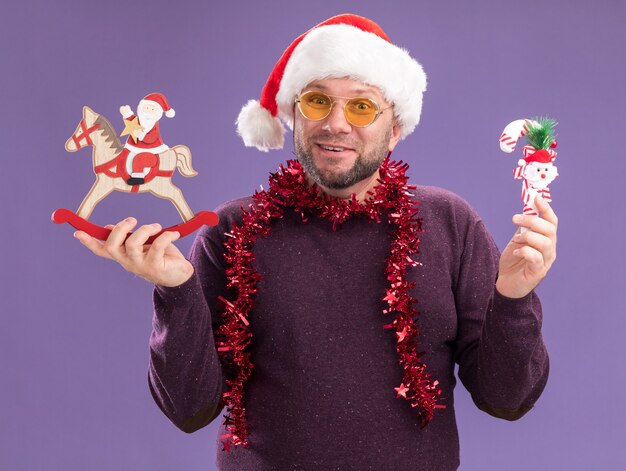 Joyeux homme d'âge moyen portant bonnet de Noel et guirlande de guirlandes autour du cou avec des lunettes tenant l'ornement de canne à sucre et le père Noël sur figurine cheval à bascule isolé sur mur violet