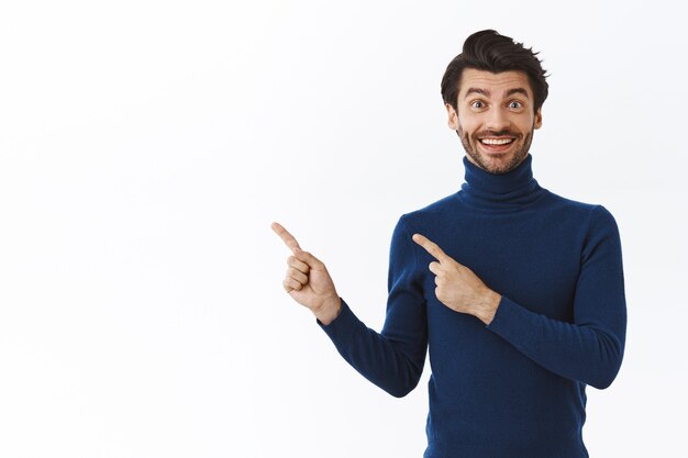 Joyeux homme barbu gai en pull bleu, recommande un produit génial, souriant impressionné, pointant le coin supérieur gauche, fait la promotion de quelque chose sur un mur blanc